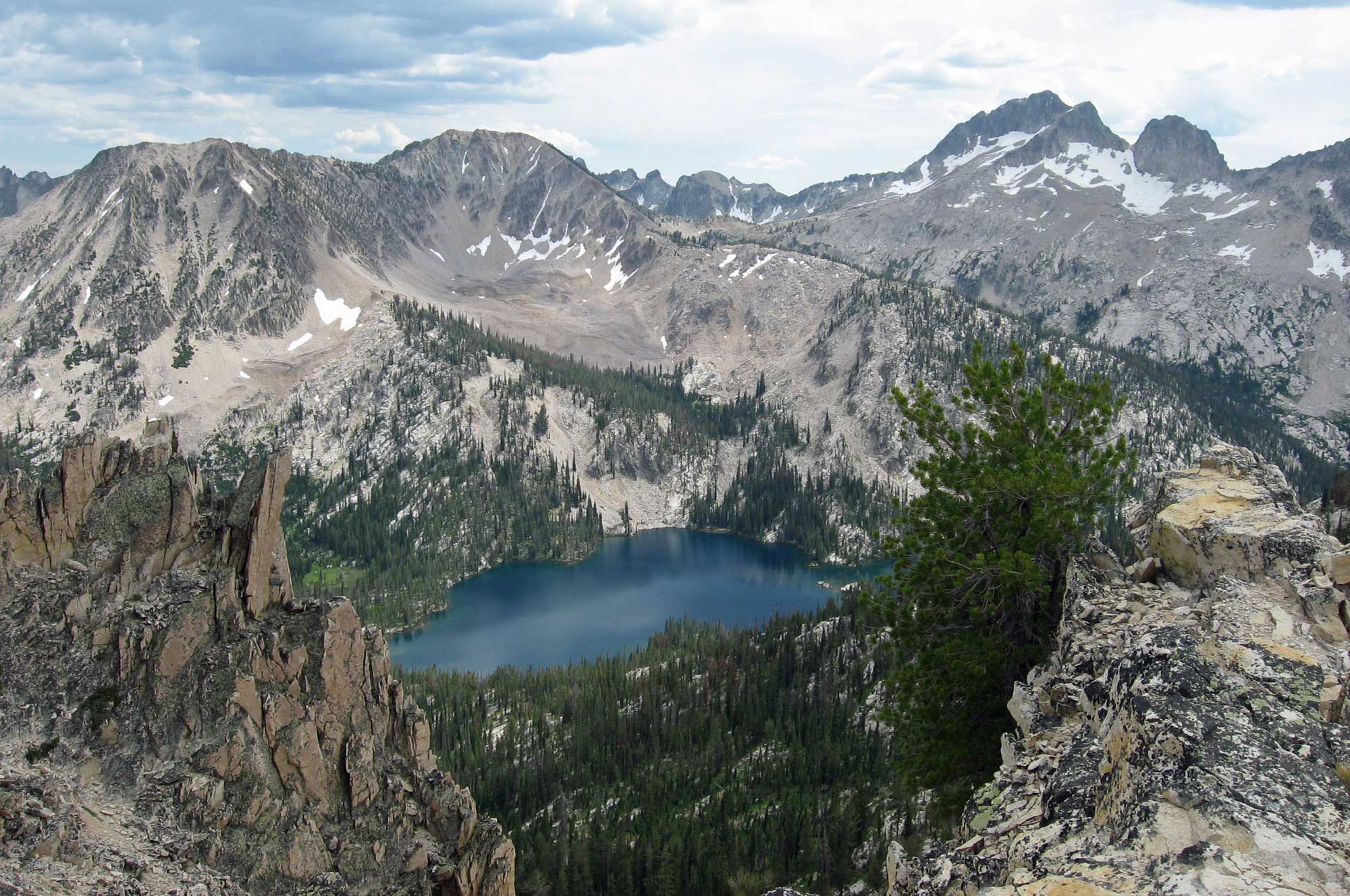 Photograph of Toxaway Lake surrounded by mountains made up of granite from the Idaho Batholith. The photo shows a blue lake surrounded by jagged gray mountain peaks. The lower slopes of the mountains have conifers on them; the upper slopes have patches of snow.