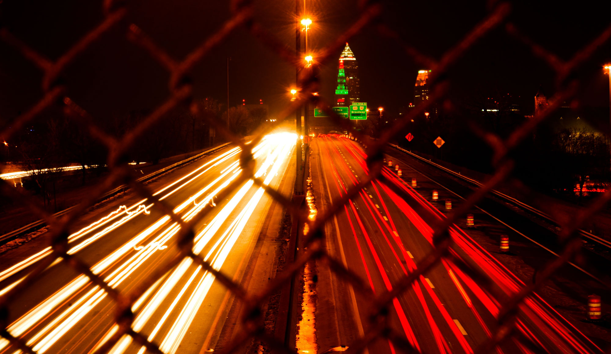 Long-exposure photograph of a divided highway in Cleveland, Ohio at night. The photo shows streaks of lights made by cars passing in two directions. White lights to the left are headlights, whereas red lights to the right are taillights. The photo was taken from an overpass and from behind a chainlink fence. Tle links, which are out of focus, can be seen in the foreground. In the background are building that are light up with yellow, green, and red lights.