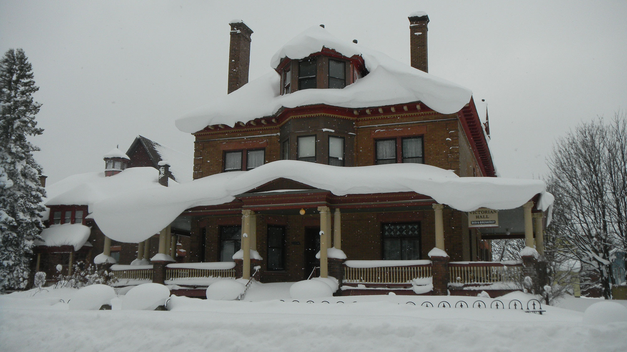 Photograph of Victorian Hall in Laurium, Michigan in the winter. The photo shows a large brick house. The roof of the house and the roof of its veranda are covered in a thick layer of snow. Snow also covers the ground around the house.
