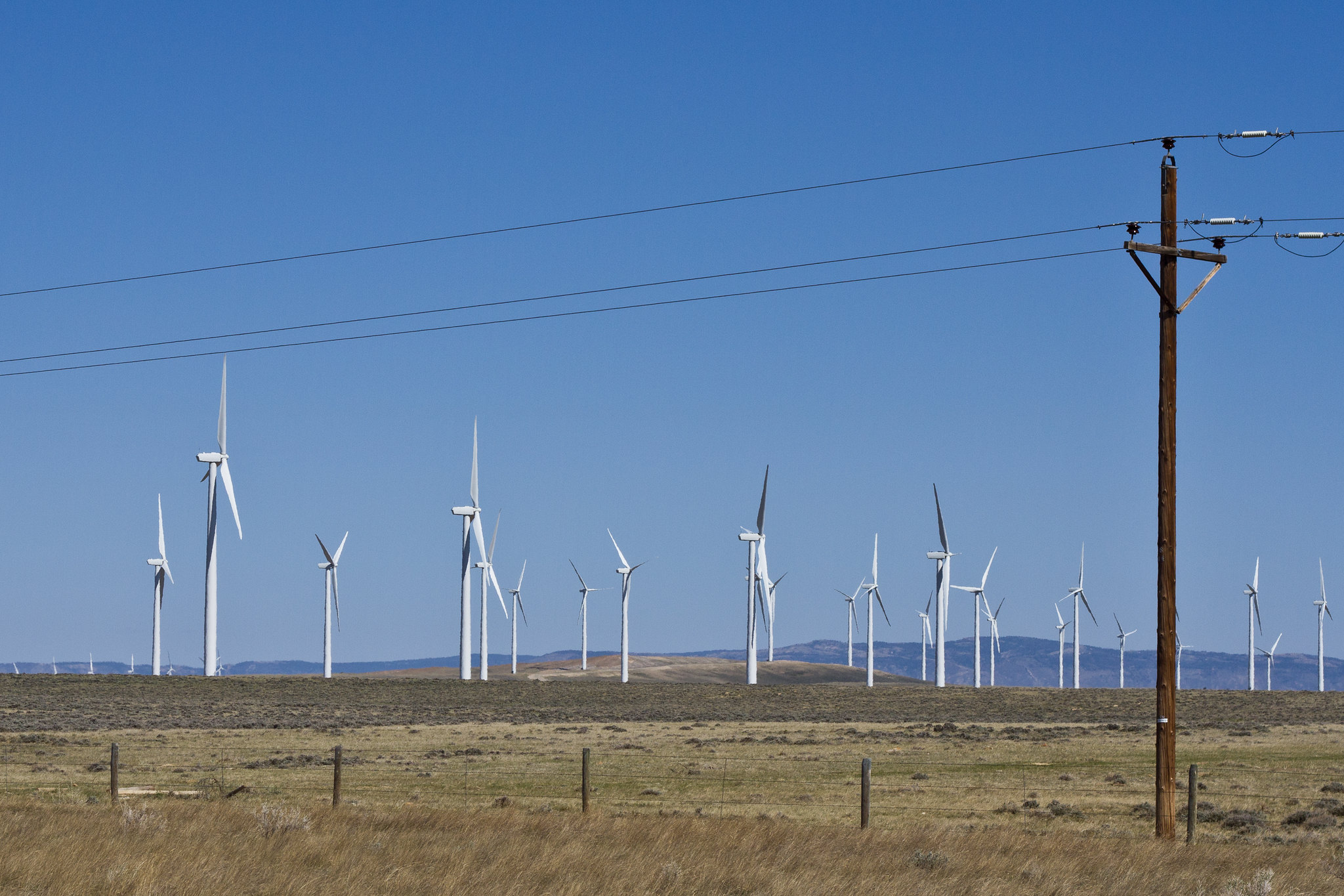 Photograph of a wind farm near Medicine Bow, Wyoming. The photo shows large white wind turbines scattered on a flat grassy and shrubby landscape. Low hills rise in the background.