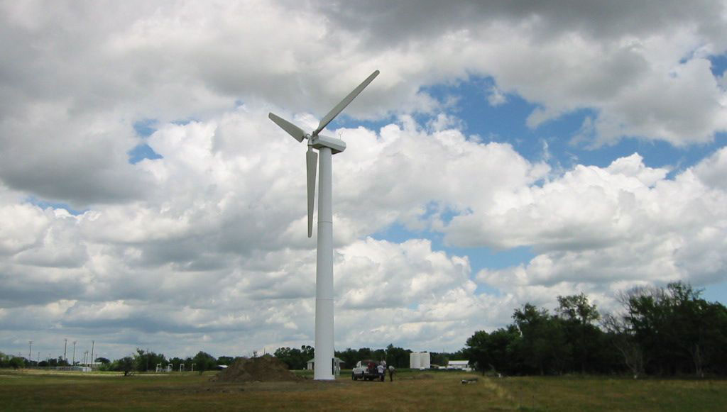 Photograph of a single wind turbine in Canova, South Dakota. The photo shows a single white wind turbine with three blades standing in a flat field. A white pick-up truck with some people near is parked at the base fo the turbine. Trees and structures are scattered in the background.