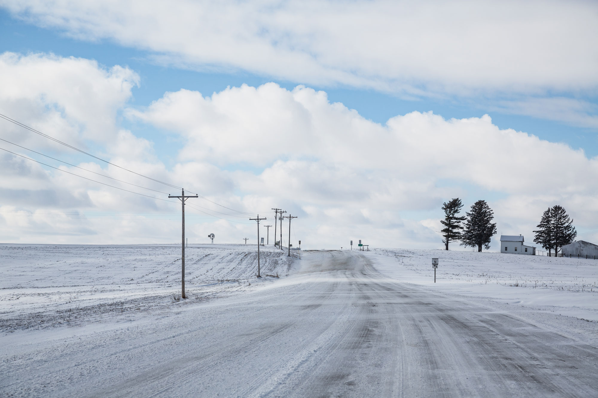 Photograph of farmland in Potosi, Wisconsin during the winter. The photo shows a road running toward the crest of a shallow hill, flanked by field on either side. On the right side of the road at the top of the hill is a house and outbuilding with several coniferous trees. A thin layer of snow covers the fields and road. White streaks crossing the road horizontally indicate that snow is blowing over the road. Power lines on wooden poles parallel the left side of the road. The sky is blue with fluffy white clouds.