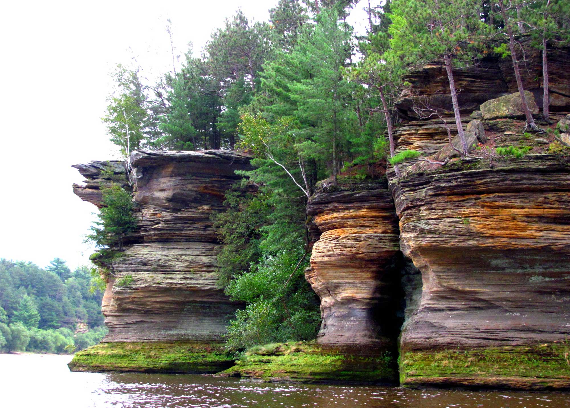 Photograph of the Wisconsin Dells. The photo shows rounded sandstone cliffs with water at their base. Trees are growing between and on top of the cliffs.