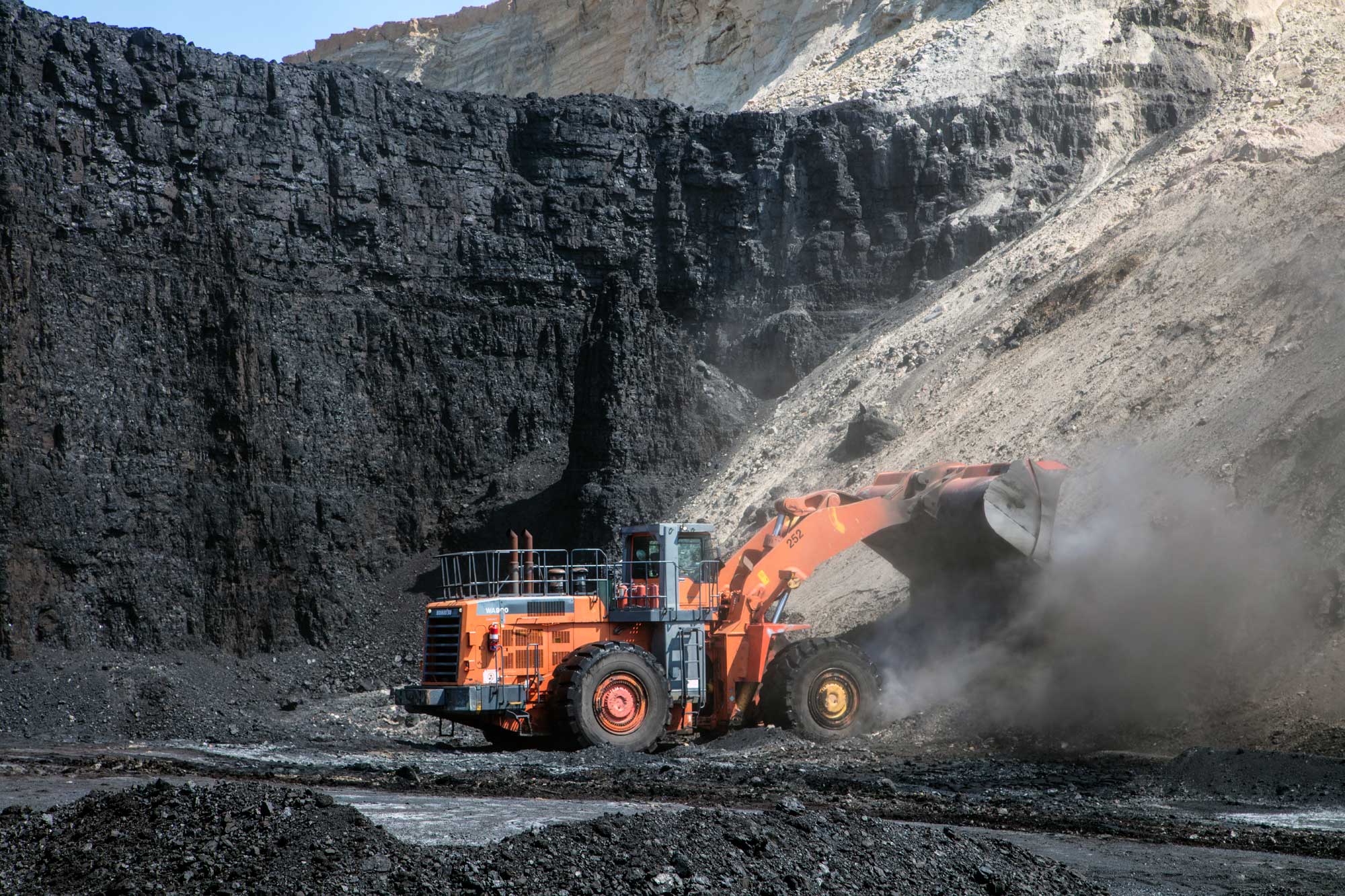 Photograph of a front-end loader at work in a coal mine. The photo shows a vehicle with a shovel attached to the front. The machine appears to be dropping a load of rock onto a pile. Next to the pile and behind the front-end loader, a thick, vertical wall of coal can be seen. The wall of coal is much taller than the machine.