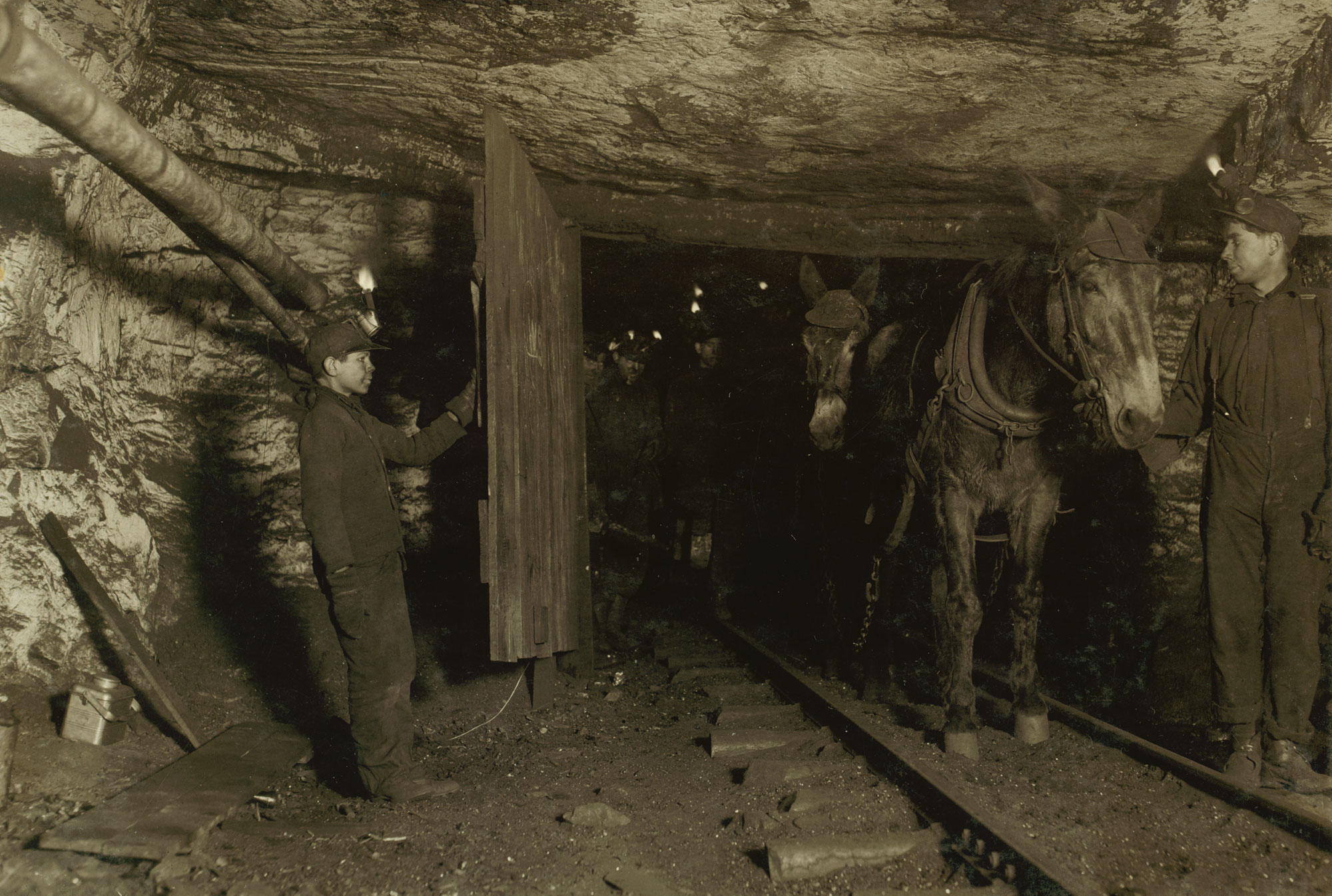 Sepia-toned photograph of boys working in a mine, 1911. The photo shows a boy of 13 years old holding open a wooden door so a mine worker (maybe a teenager) can lead mules through the door on a railway track. The boy holding the door is dressed in a dark colored coat and long pants. He is wearing a cap with a lamp on the front. The other miner is dressed similarly. At least two more miners can be seen in the gloom beyond the door. The walls and ceiling around the door are painted white.