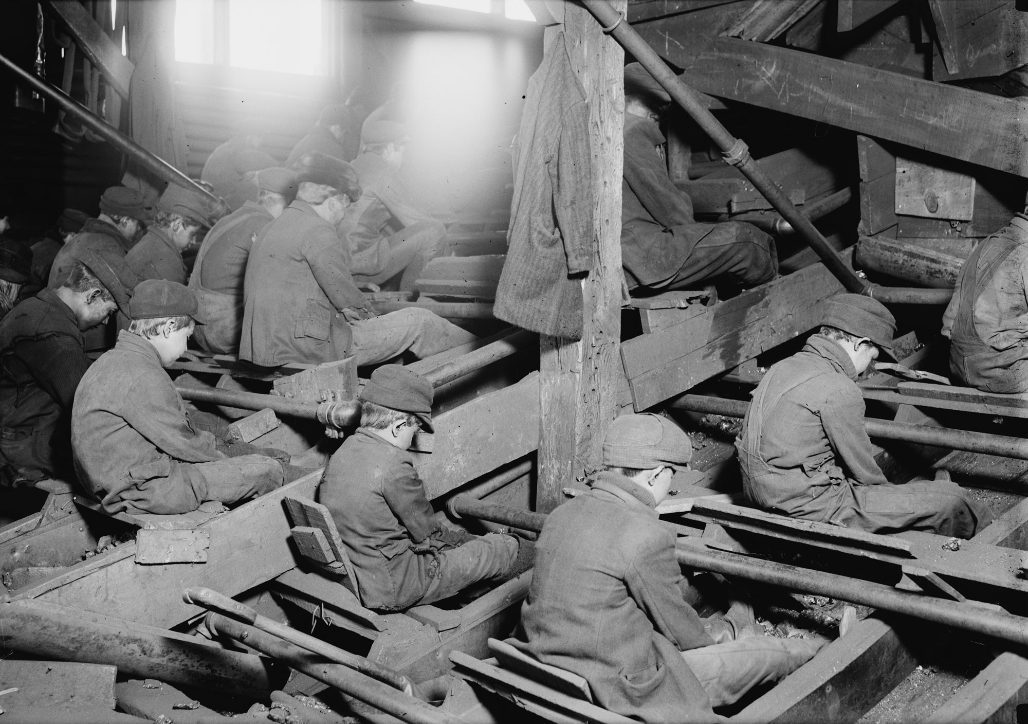 Black and white photograph of breaker boys, 1911. The photo shows rows of young boys dressed in jackets and pants, each wearing a cap. The boys each sit in a seat. The boys are each looking down into a trough at their feet. Light shines through windows in the background.