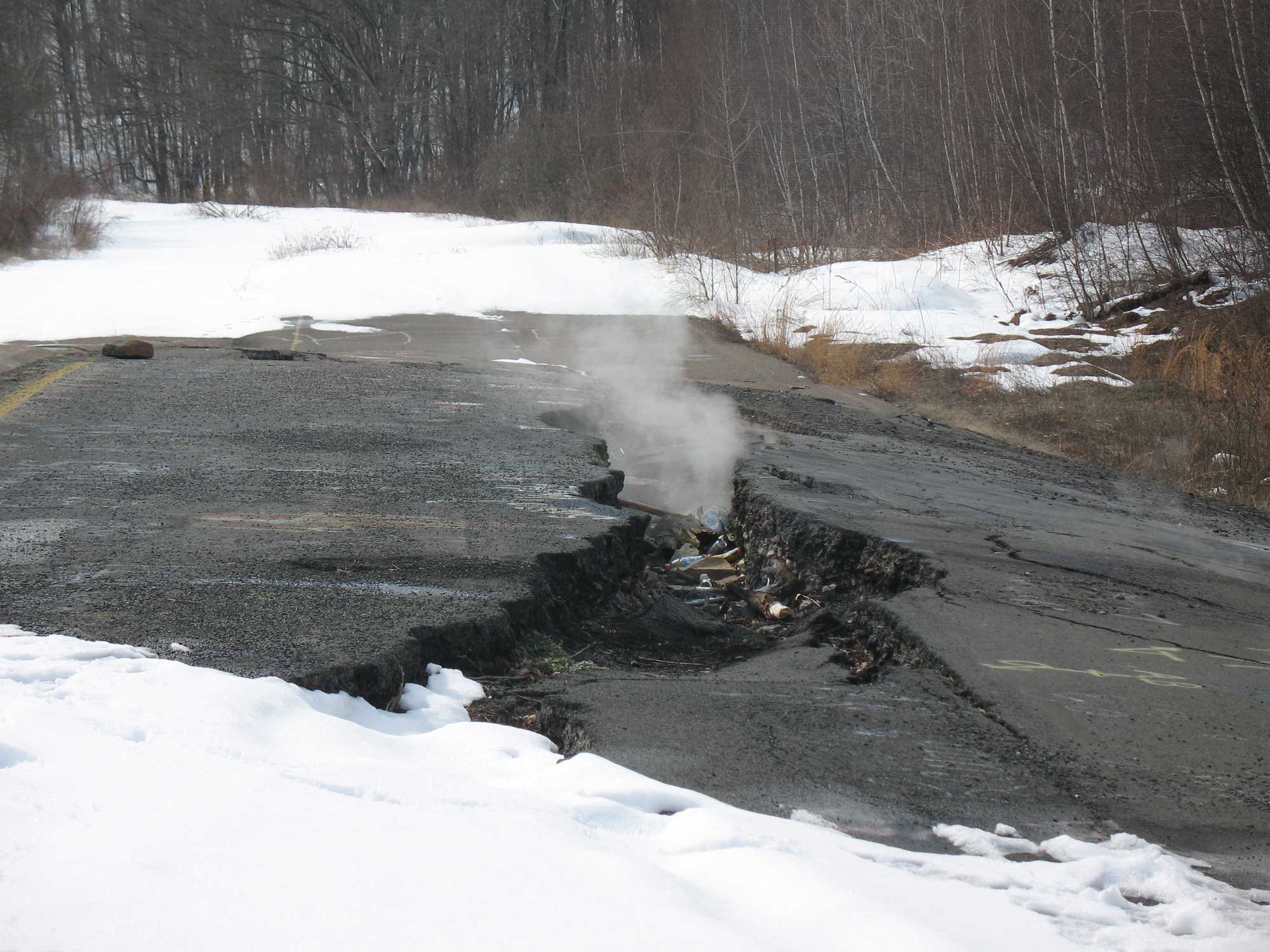Photograph of the effects of the Centralia, Pennsylvania, underground mine fire from 2010. The photo shows an asphalt road with fissure in it and smoke coming out of the fissure. While the area immediately around the fissure has no snow, snow can be seen in the foreground and background of the image.