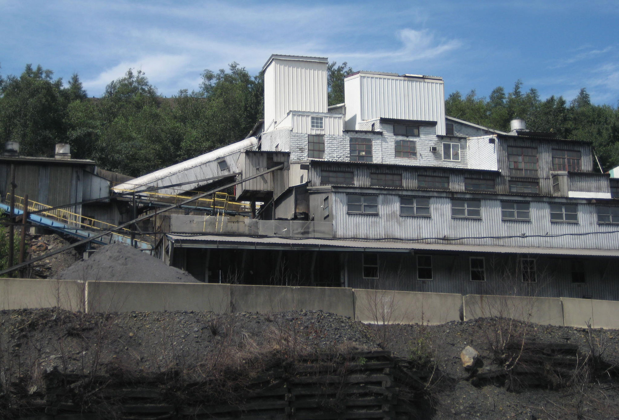 Photograph of a modern coal breaker plant in Pennsylvania. The photo shows a building with white paneling behind a concrete barrier. A chute emerges from the left side of the building.
