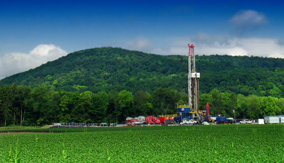 Photograph of a gas drill rig in Pennsylvania. The photo shows a drill rig, vehicles, and equipment in the mid distance. In the foreground is a green field. In the background is a forested hill. The drill rig looks like a thin, rectangular scaffold tower with a blue and yellow platform at its base. 