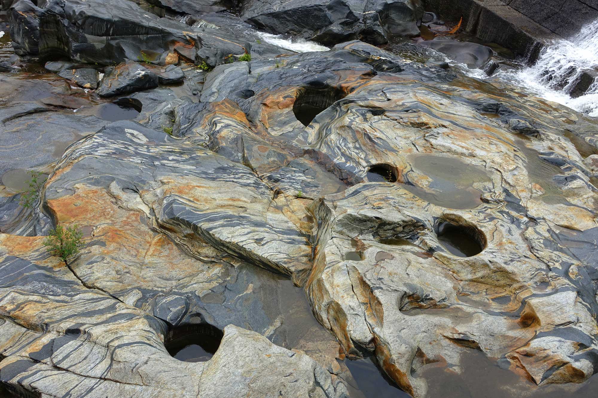 Photograph of potholes at Shelburne Falls, Massachusetts. The photo shows beige rocks with swirling gray stripes and areas stained orange. The rock has a number of potholes, or small circular depressions, in it. Water can be seen flowing over a barrier in the upper right corner of the image.