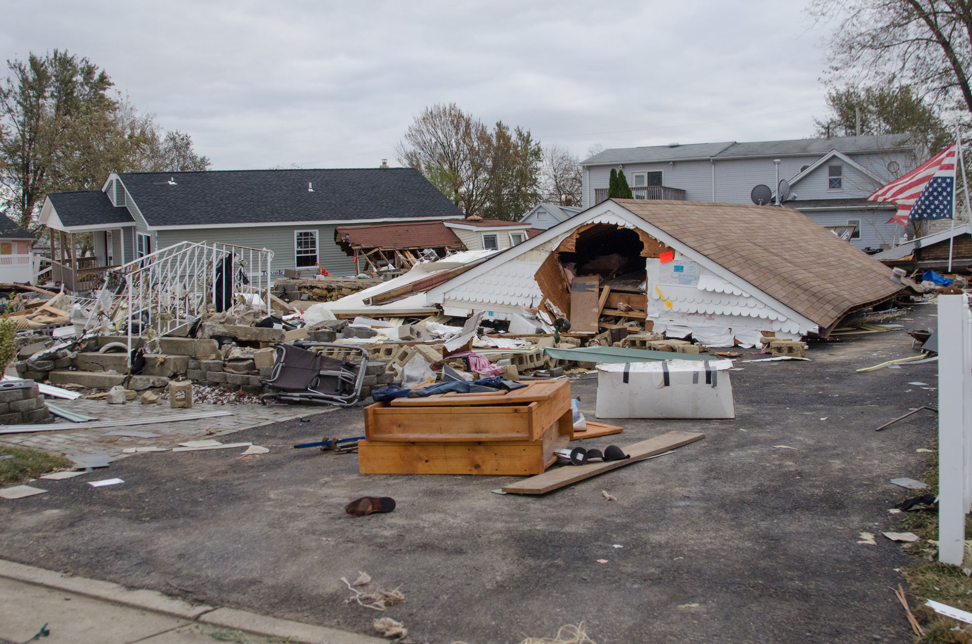 Photograph of damage in Union Beach, New Jersey, following Hurricane Sandy in 2012. The photo shows a house mostly swept away by the storm, leaving only a roof sitting at the end of the driveway. Stairs that formerly led to the house can be seen at left, and debris is strewn around. Other houses that were apparently less damaged stand in the background.