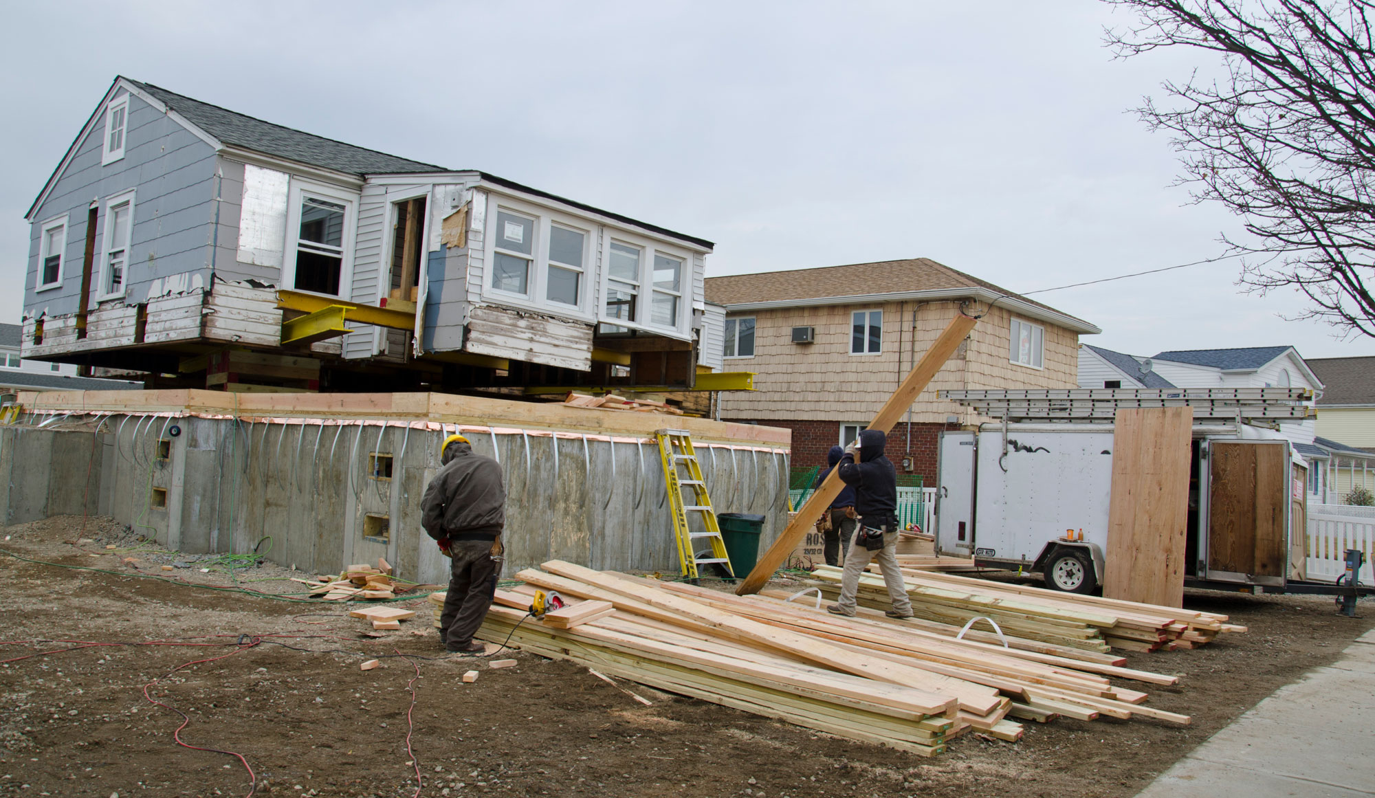 Photo of a house being raised following Hurricane Sandy. The photo shows a gray single-family home on a cement pedestal. The house appears to be sitting on blocks or beams. In the foreground are two workers, a work truck, and a pile of boards. More houses extend away from the viewer toward the right.