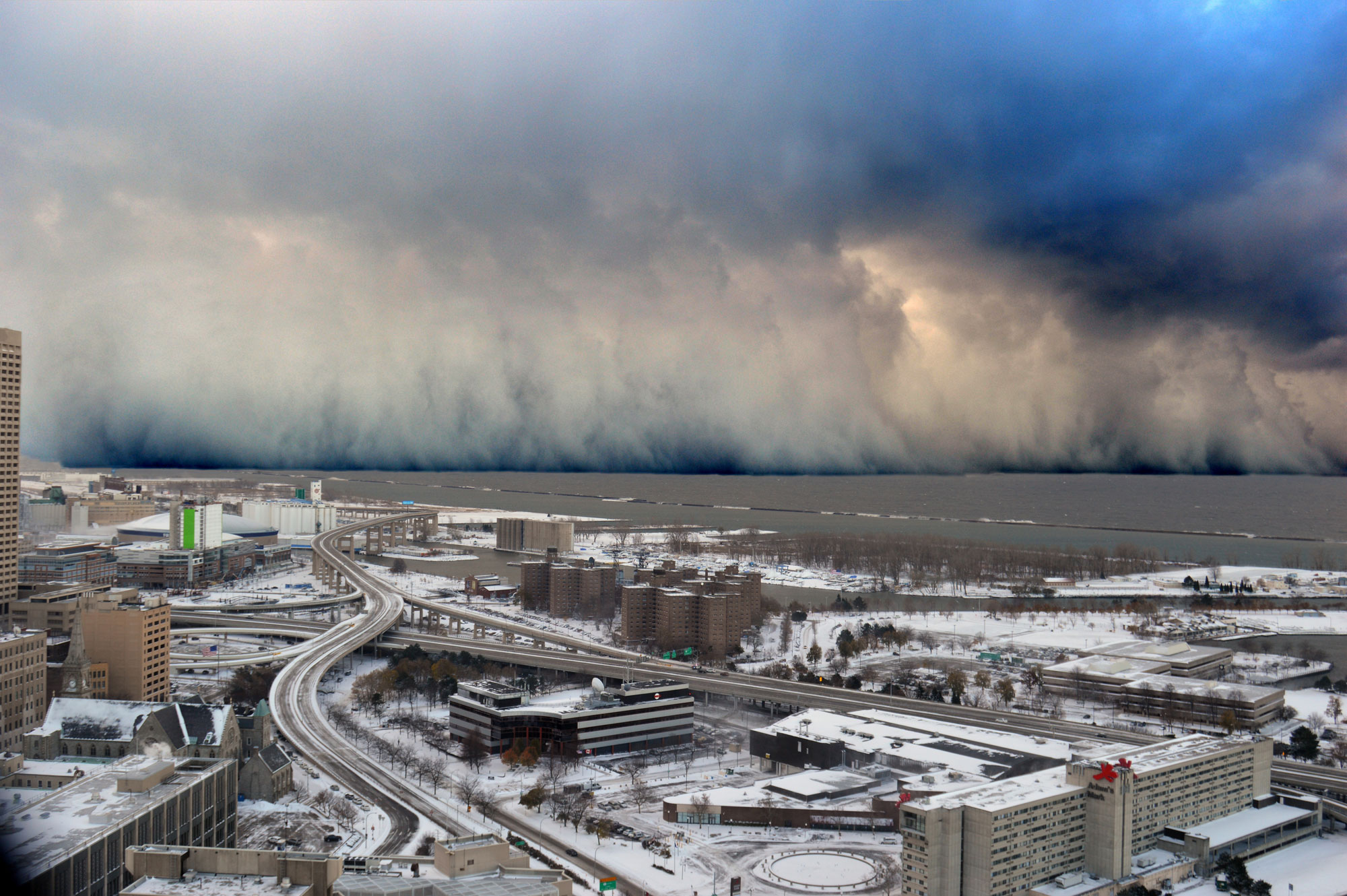 Photo of a cloud of lake effect snow bearing down on Buffalo Southtowns, New York, in 2014. The photo shows part of a city in the foreground, and the shore of Lake Erie in the mid-distance. Clouds extend from the surface of the lake upwards to a ceiling of clouds in the distance.