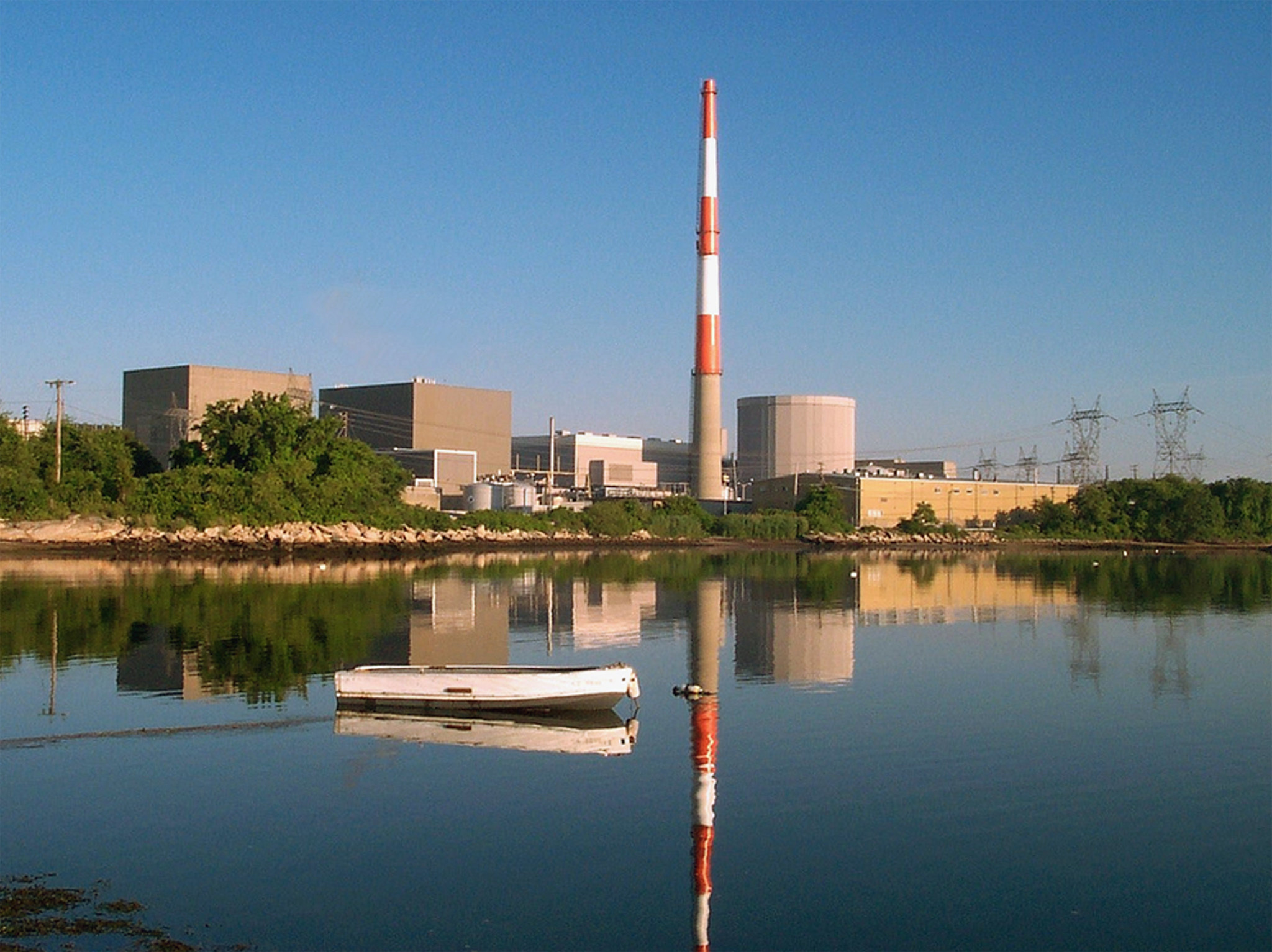 Photograph of the Millstone Power Station, Waterford, Connecticut. The photo shows rectangular buildings, a cylindrical building, and a red and white striped smokestack on the shore net to a body of water.