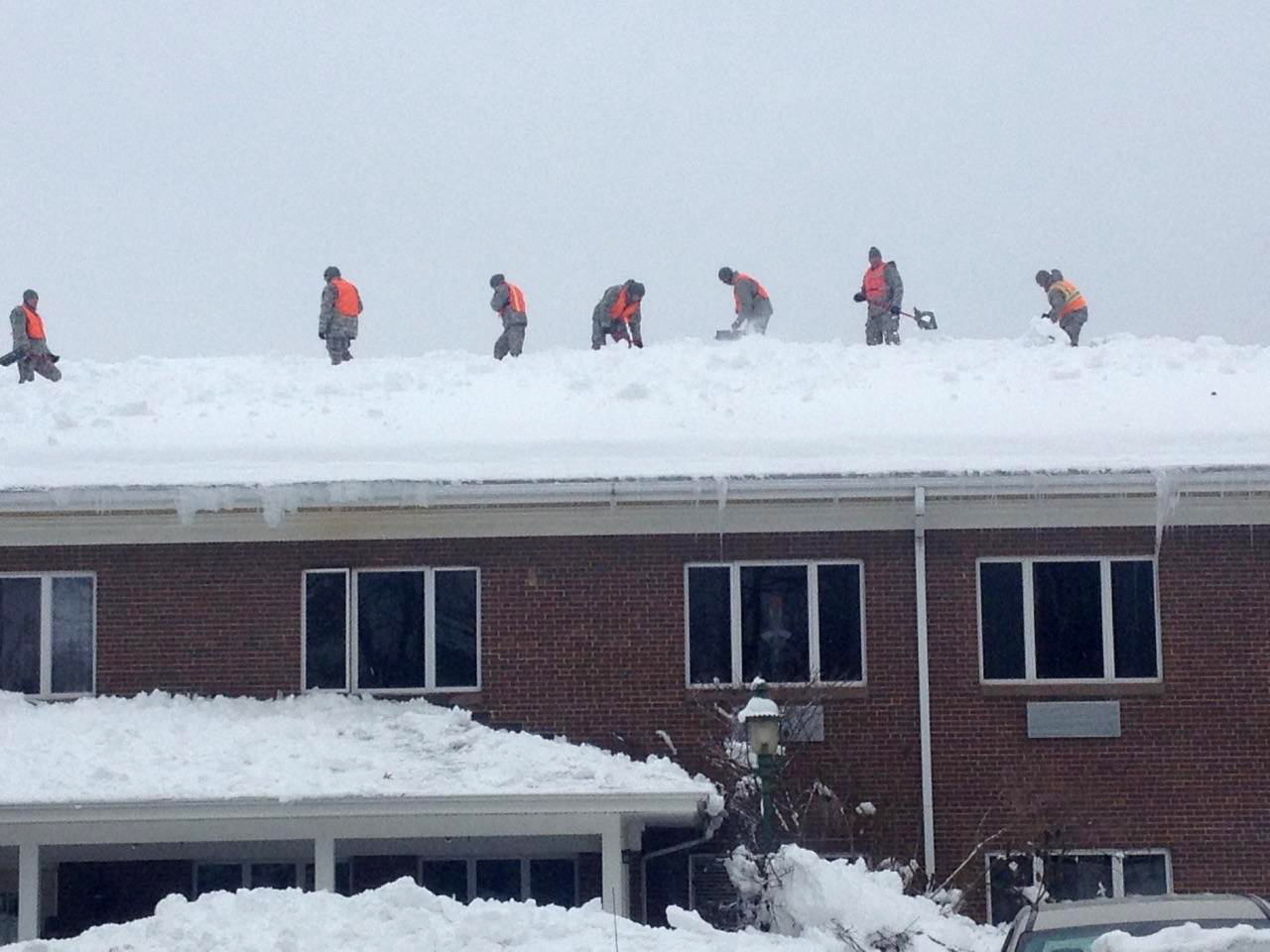 Photograph of National Guard members on the roof of a two-story brick building shoveling snow. The photo shows seven guard members in fatigues and orange vests standing on the roof, about five of whom clearly have snow shovels. 
