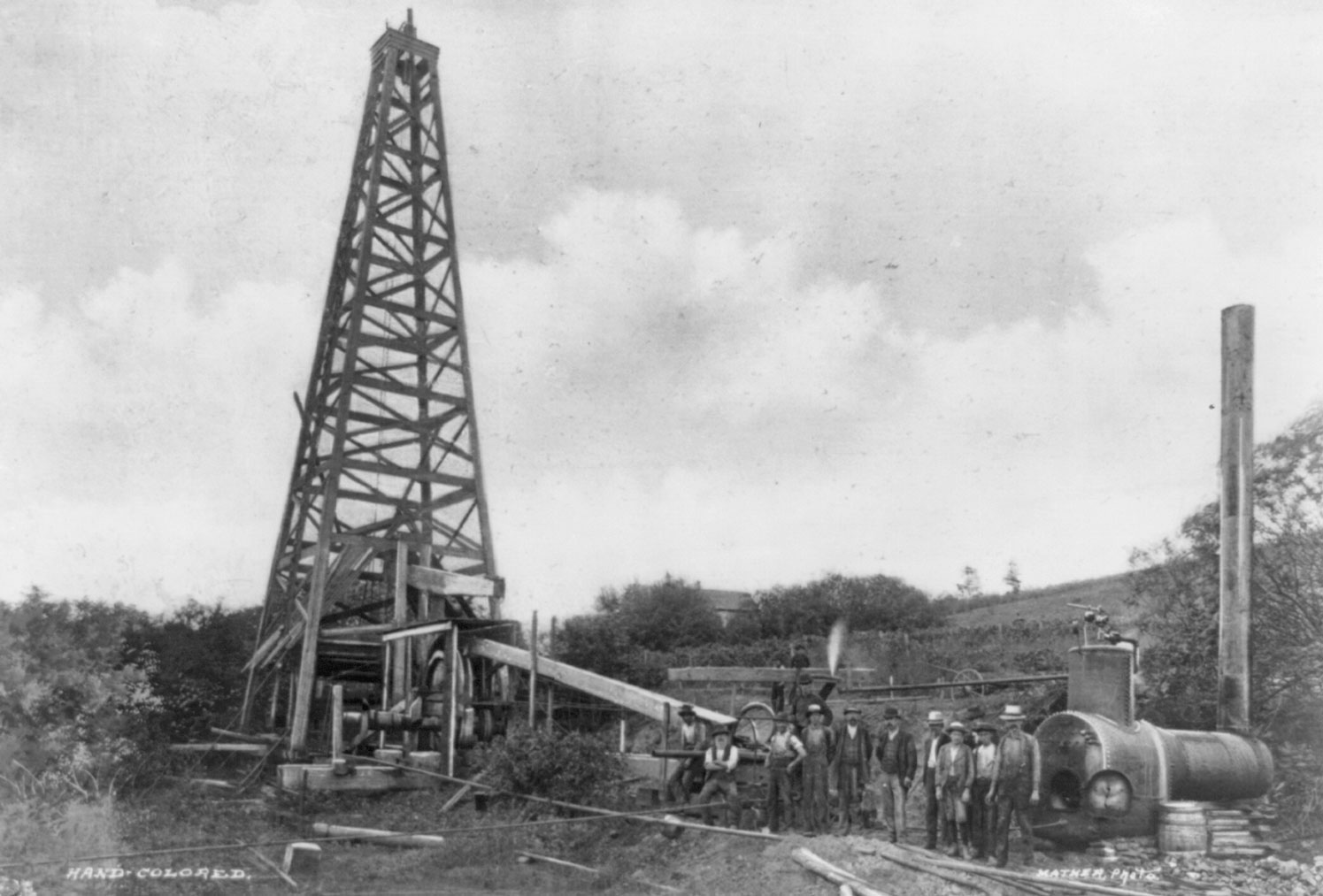 Black and white photograph of an oil derrick in Titusville, Pennsylvania, around 1900. The photo shows a derrick, which looks like a taper tower made out of wood or metal beams, rising to the left. In the right foreground, a group of men stands. Other equipment is scattered behind and around the men.