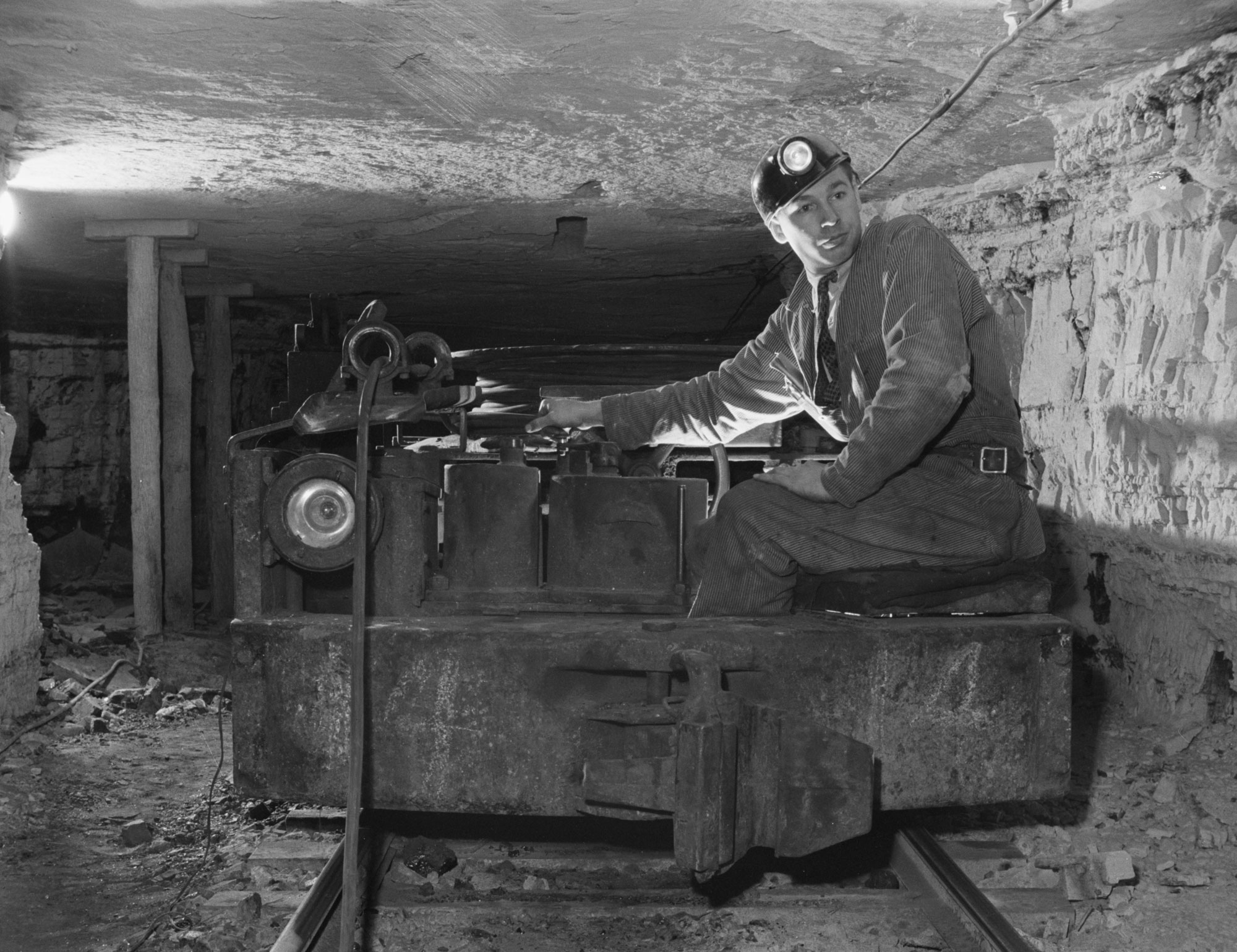 Black and white photograph of a man operating a mine car, 1942. The photo shows a man in coveralls over a shirt and tie, with a hard hat that has a lamp on the front. The man is seated on the right side of a mien car, a low car that runs on rails. The man and car are in a mien shaft with a low ceiling. In the left background, three supports can be seen holding up the ceiling.