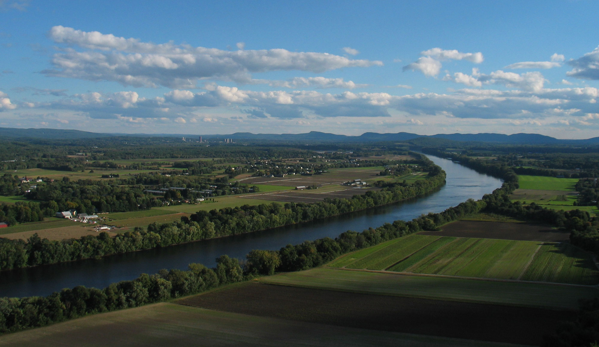 Photograph of the Connecticut River Valley (also known as Pioneer Valley) during the summer. The photo shows a river beginning at the lower left of the image and proceeding diagonally upwards and to the right, bending near the right side of the image and proceeding toward the horizon. The area on either side of the river is flat with patches of forest and agricultural fields. The Holyoke Mountains rise in the far distance.