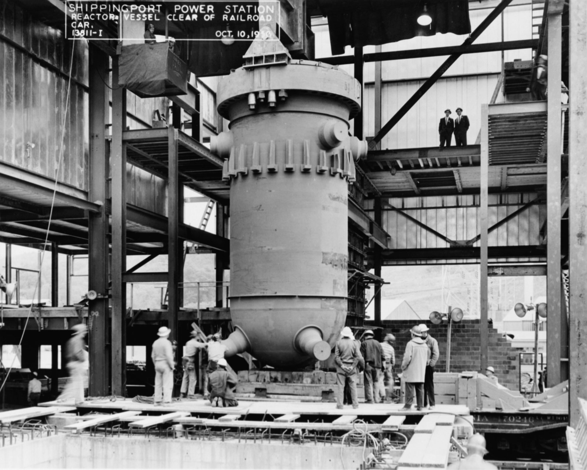 Black and white photograph of a reactor vessel for the Shippingport Atomic Power Station in 1956. The photo shows a tall cylindrical vessel sitting on a wooden pallet. A group of men stand around the base of the vessel. A platform partially wraps around the top of the vessel, and there are two men standing on it looking down.