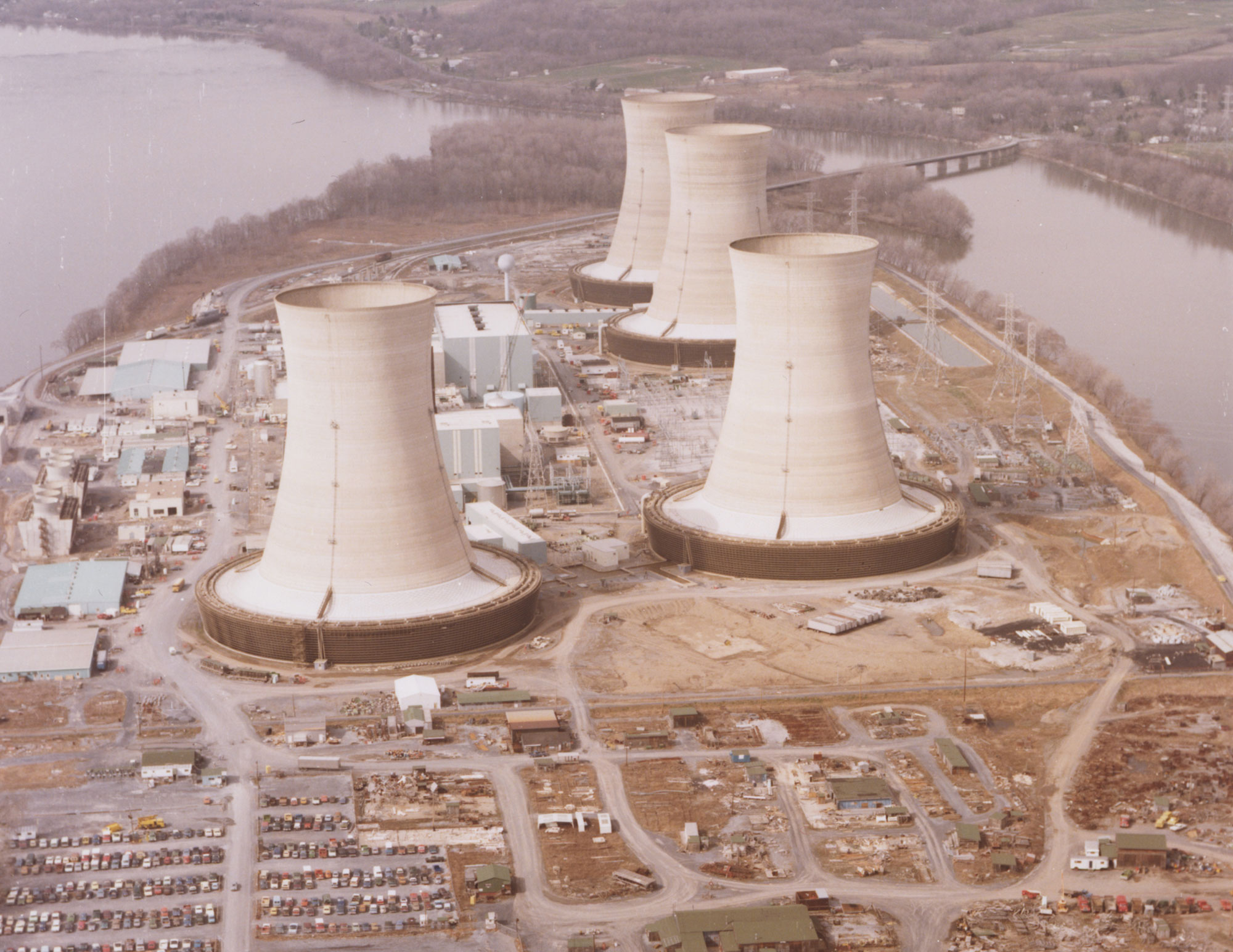 Aerial photograph of Three Mile Island Nuclear Station in 1979. The photo shows four cooling towers and associated structures on a point of land surrounded by water in the background.