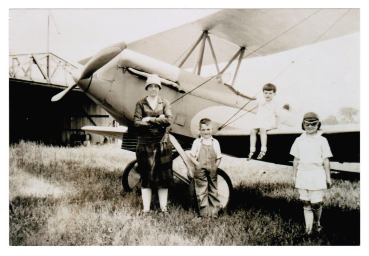 Photograph from the early 20th century of a woman and three children by an airplane on the ground.