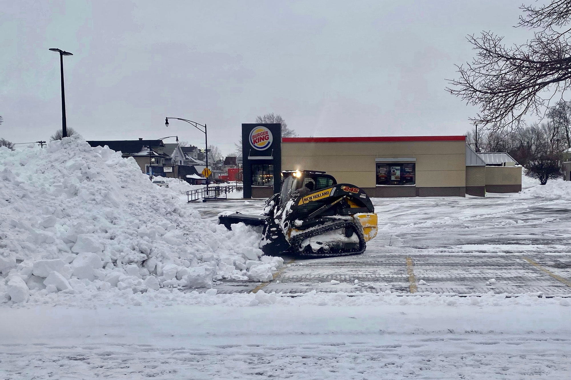 Photograph showing a small bulldozer clearing snow from the parking lot of a Burger King restaurant following a large snowstorm in Buffalo, New York, in December 2022. The photo shows a small black and yellow bulldozer pushing snow into a big pile. The restaurant can be seen in the background.