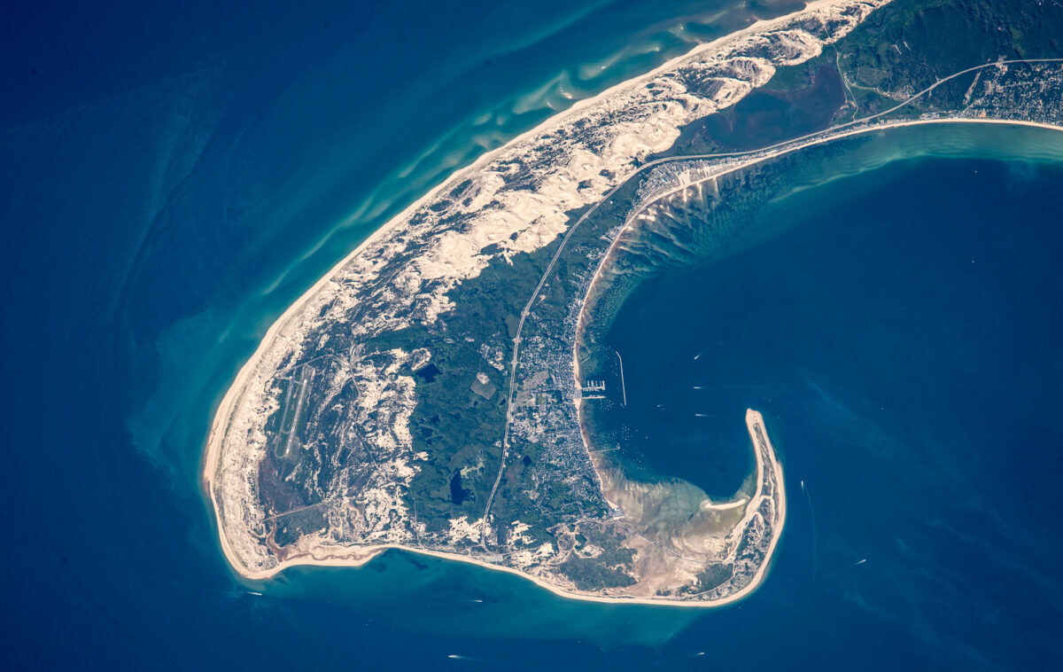 Photograph of the tip of Cape Cod, Massachusetts, taken from space. The photo shows the hook-like tip of Cape Cod, with looks sandy on its south shore (on the left side in this image) and covered with vegetation on its north shore (on the right side of this image).