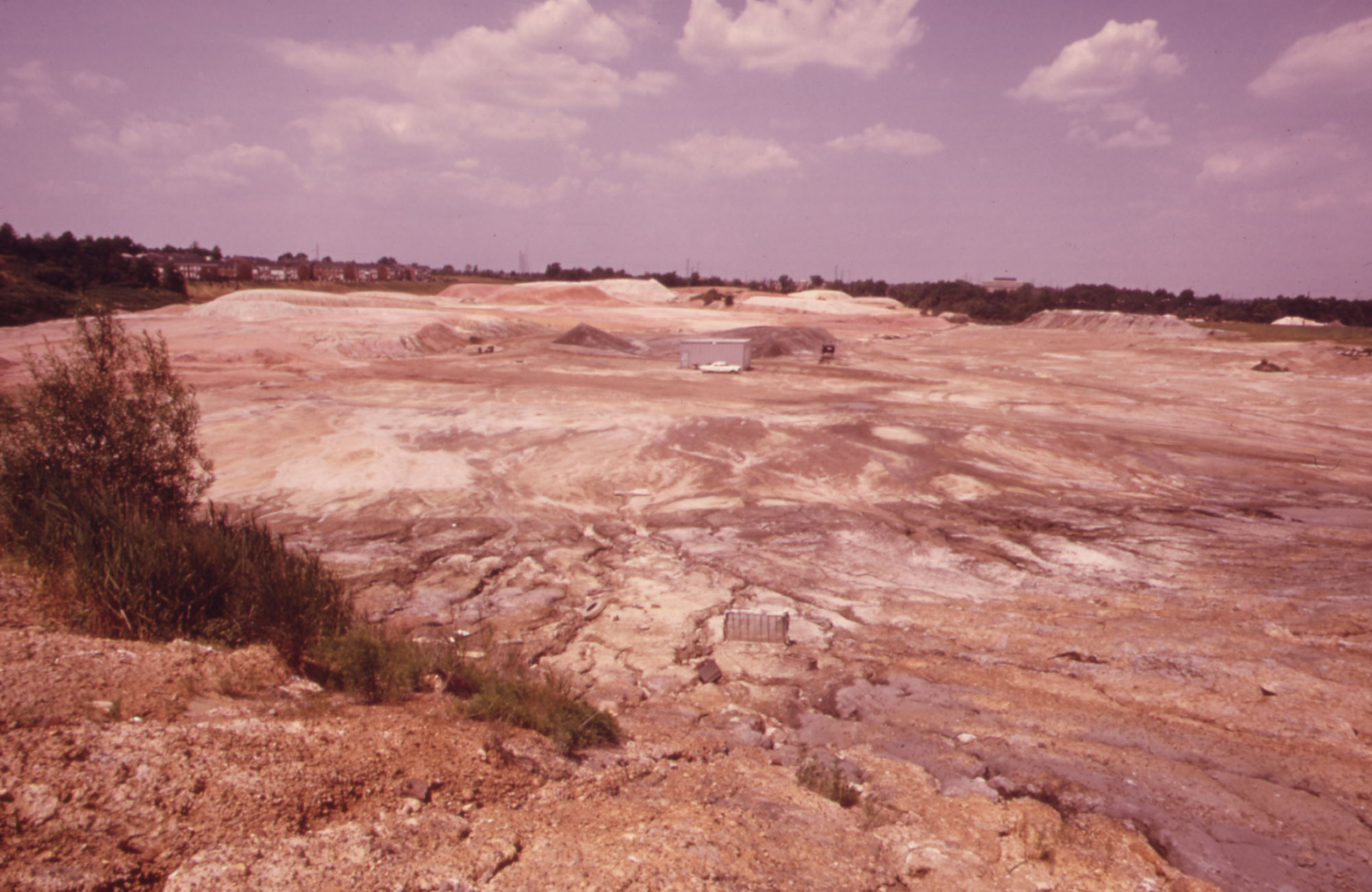 Photograph of a clay pit in Woodbridge, New Jersey, taken in 1973. The photo shows a large, mostly flat expanse of exposed clay with plants and buildings around the edges.