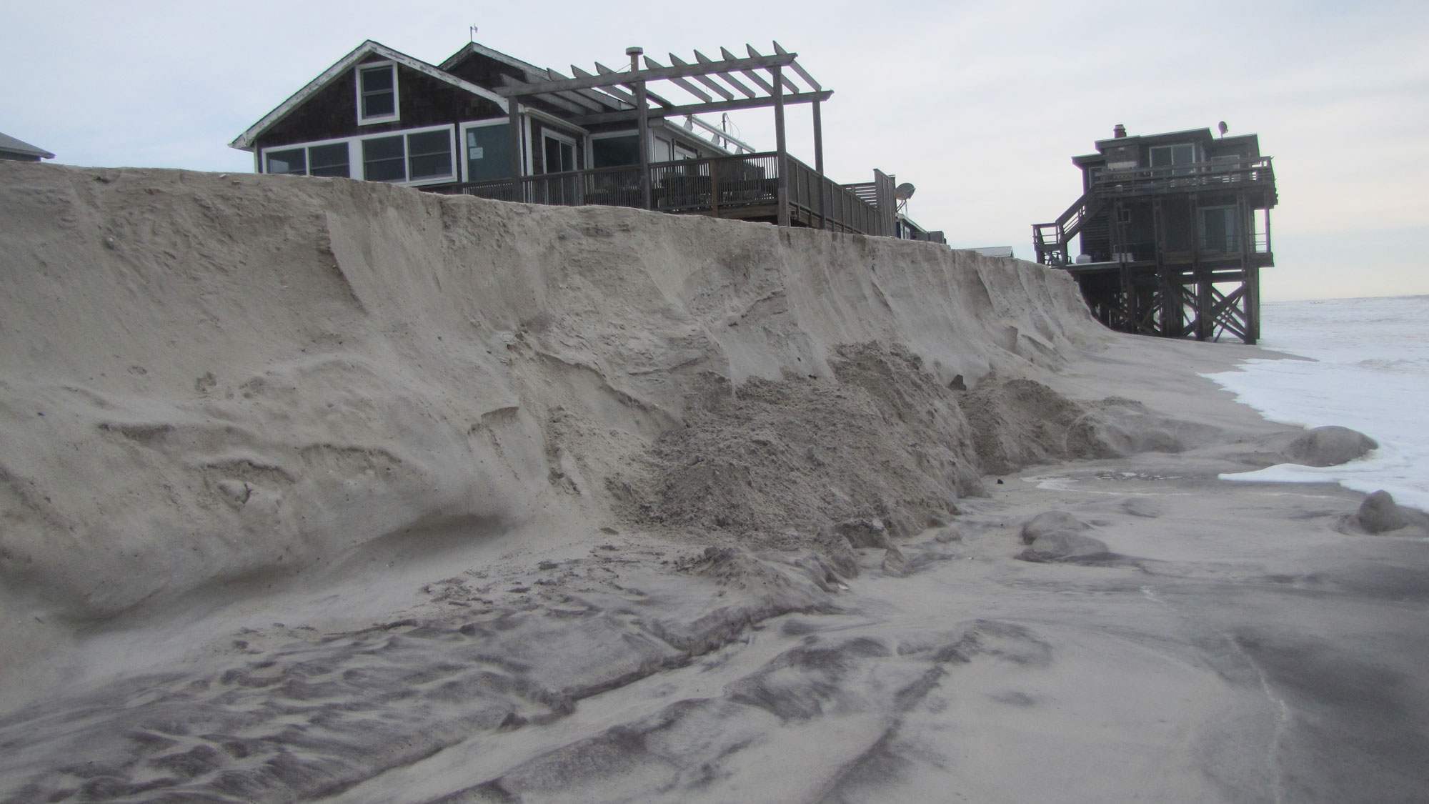 Photograph of erosion of an artificial dune on Fire Island, New York, following hurricane Sandy. The photo shows a dune with a shear face. A house sits on top of dune. In the background, another house stands at the water's edge; it is built on tall scaffolding so that the structure is above water level.