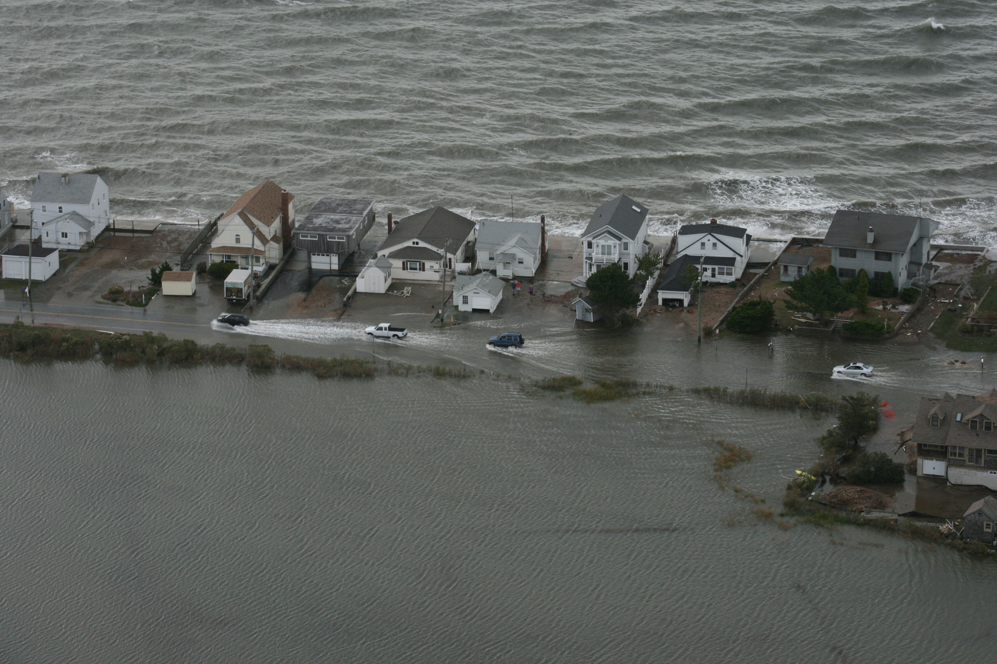 Aerial photograph of flooding in Connecticut following Hurricane Sandy in 2012. The photo shows a line of houses along a street. The landward area behind the houses is completely flooded, and the opposite sides of the houses face the ocean. Cars are driving on the street, which is also flooded.