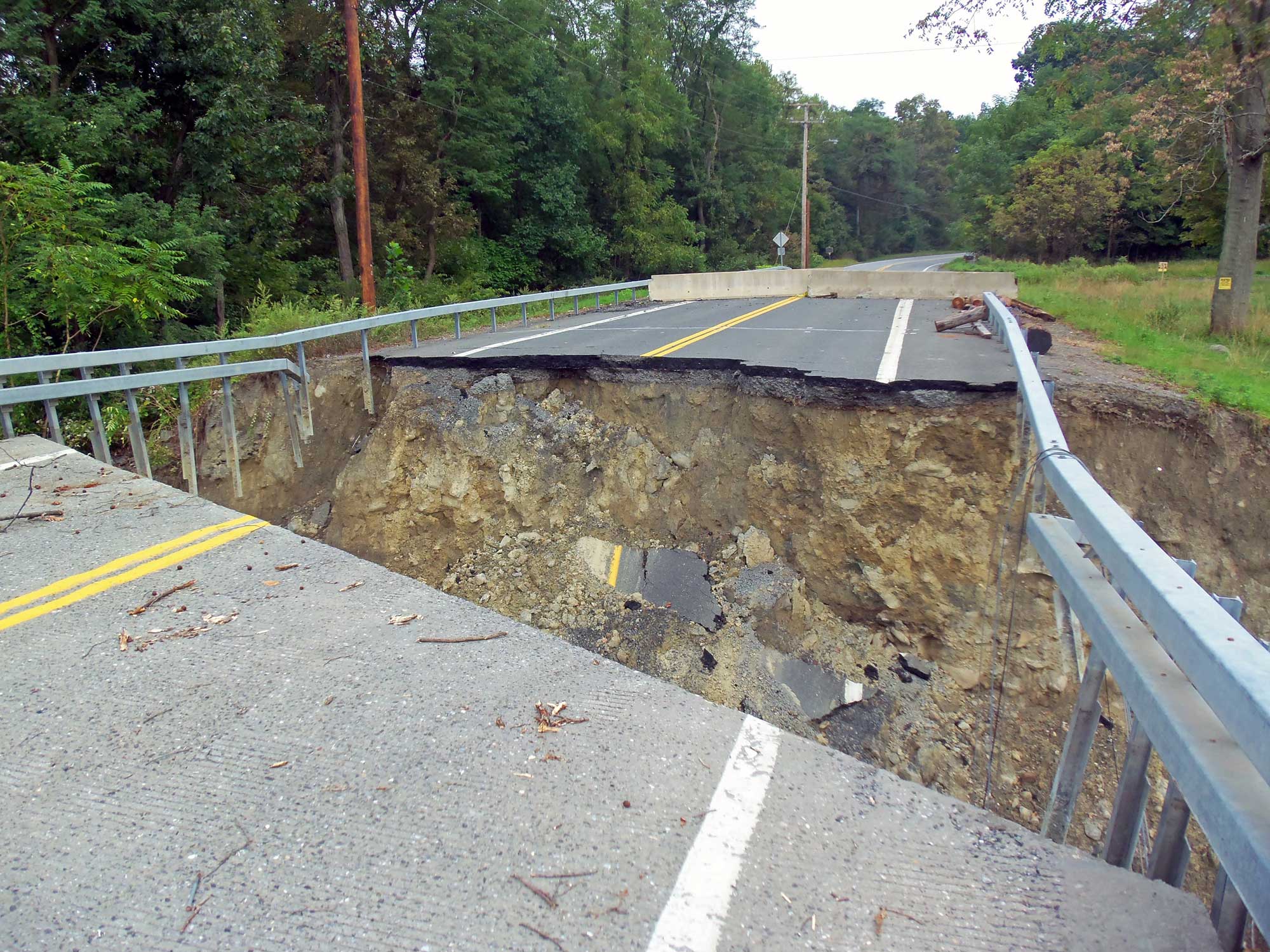 Photograph of a bridge that was washed out by flooding in Moodna Creek, New York. The photo shows a two-lane paved road with a segment missing. The photo is looking from one edge of the missing segment to the other. Dirt that supports the road can be seen on under the other side of the missing segment.