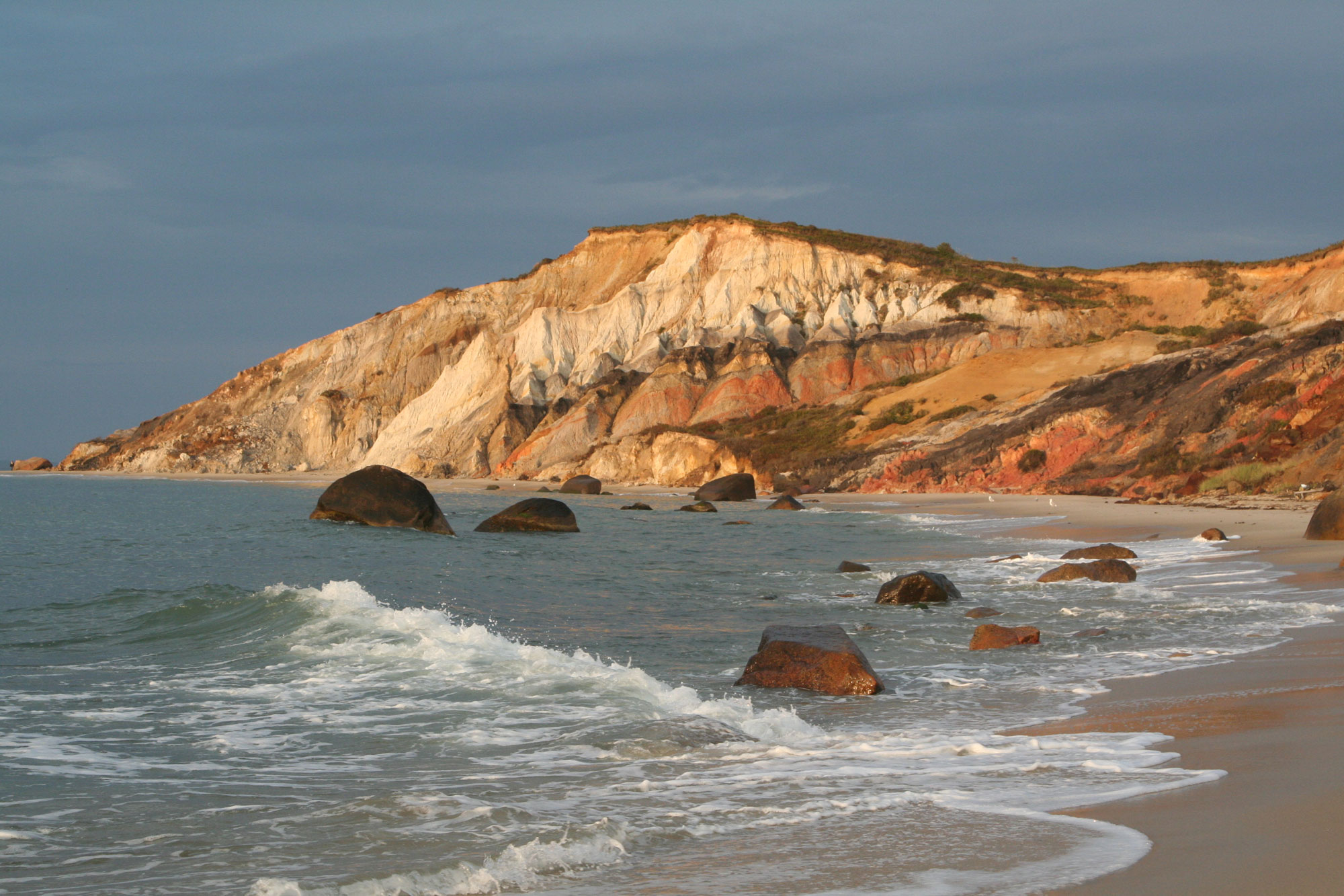 Photograph of Gay Head Cliff on Martha's Vineyard in Massachusetts. The photo shows a cliff face above a narrow beach bordering the water. The cliff is banded with red, black, and tan rocks. 