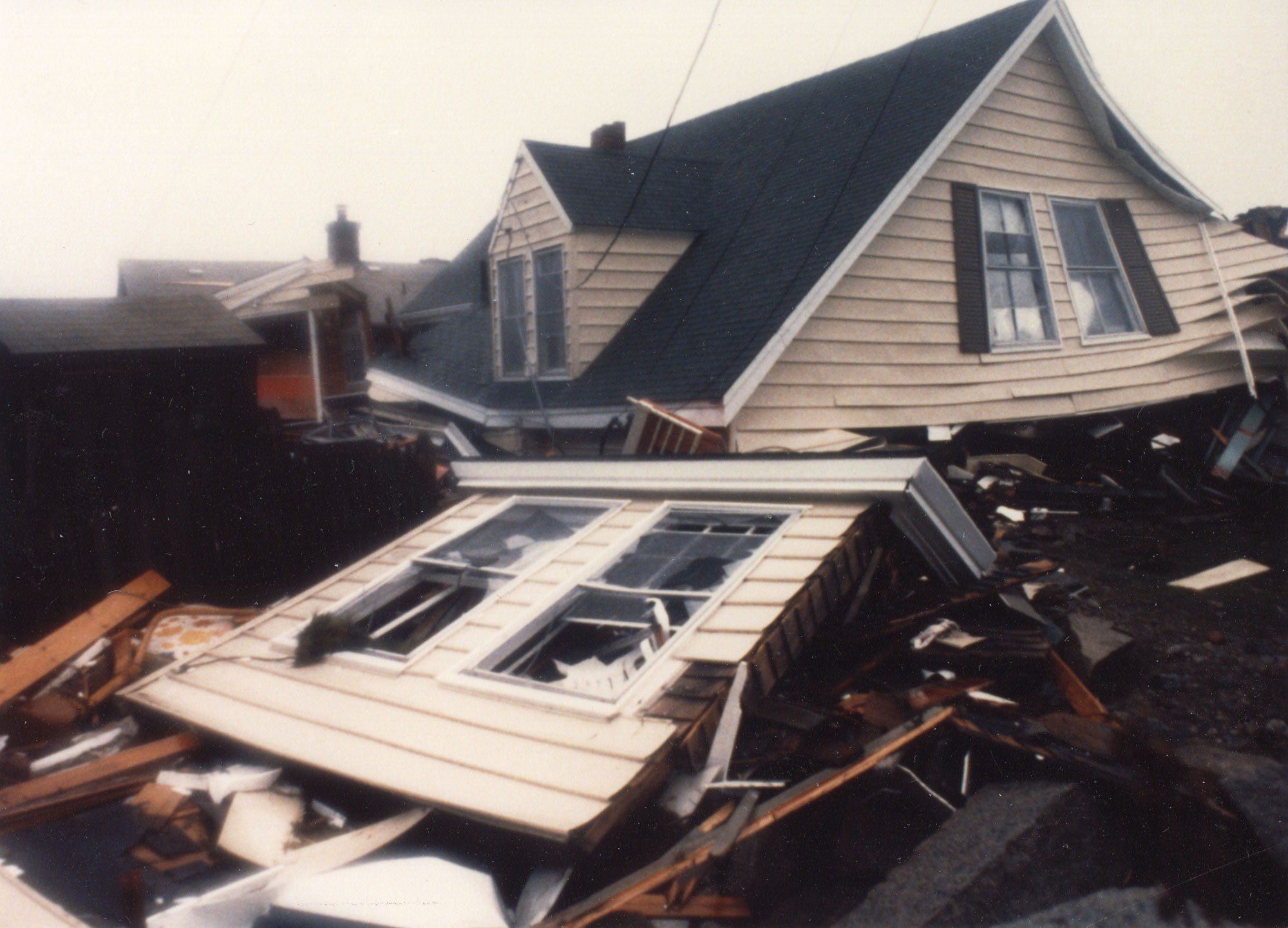 Photograph of a house in Massachusetts that was damaged during a storm in 1991. The photograph shows a house with beige or light yellow siding and a black roof. The ground floor of the house has been completely destroyed, and the roof is lying on a pile of debris.