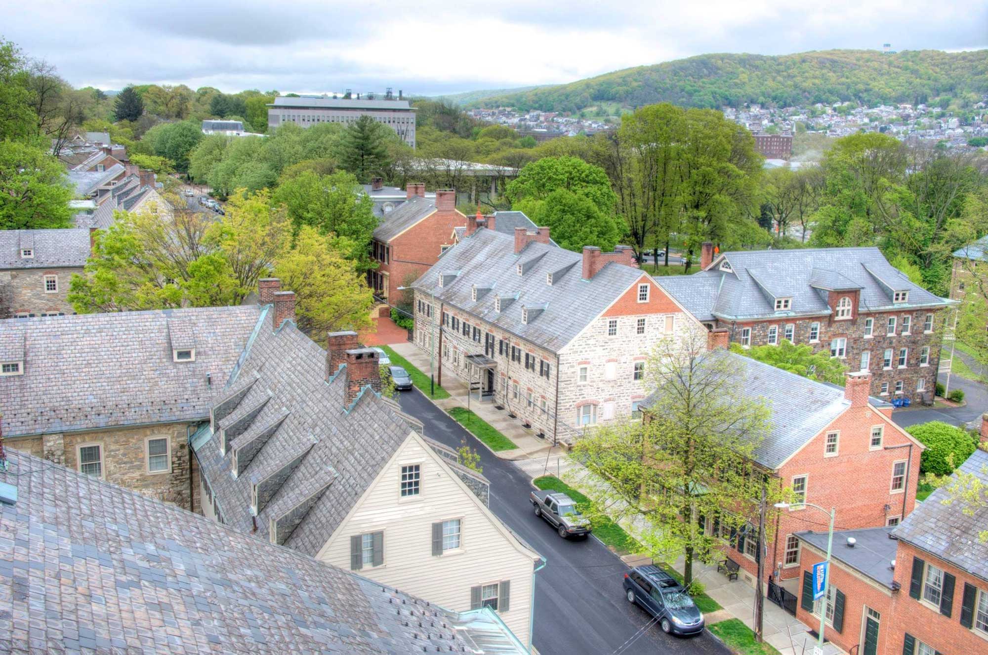 Photograph of a historic area of Bethlehem, Pennsylvania, take from a high point. The photo shows old brick buildings along a paved street, with tree-covered hills and more buildings in the background.