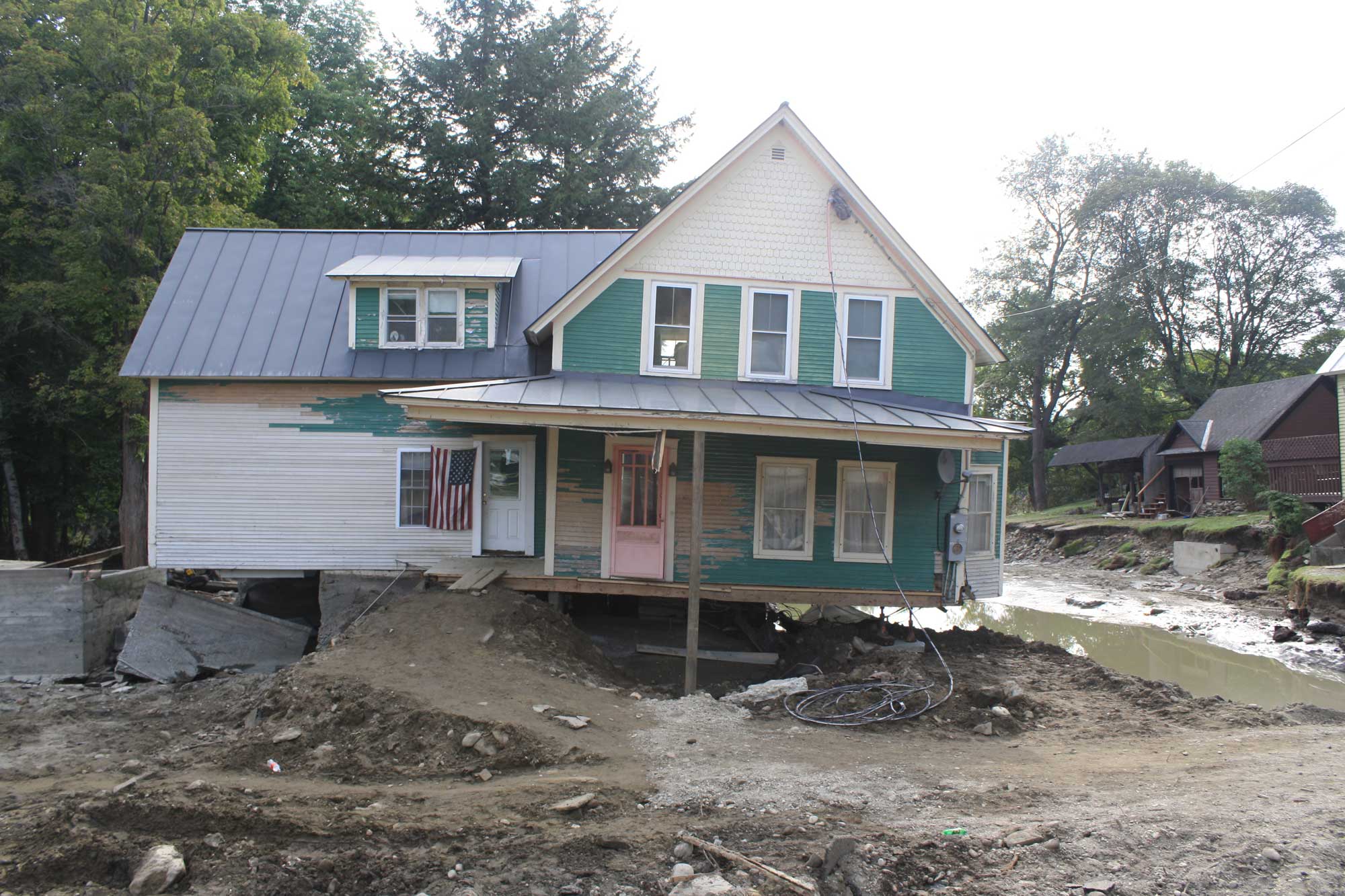 Photograph of flood damage in the town of Bethel, Vermont. The photo shows and white and green house from which much of the ground underneath was washed away. Other erosion to the landscape can be seen in the background.