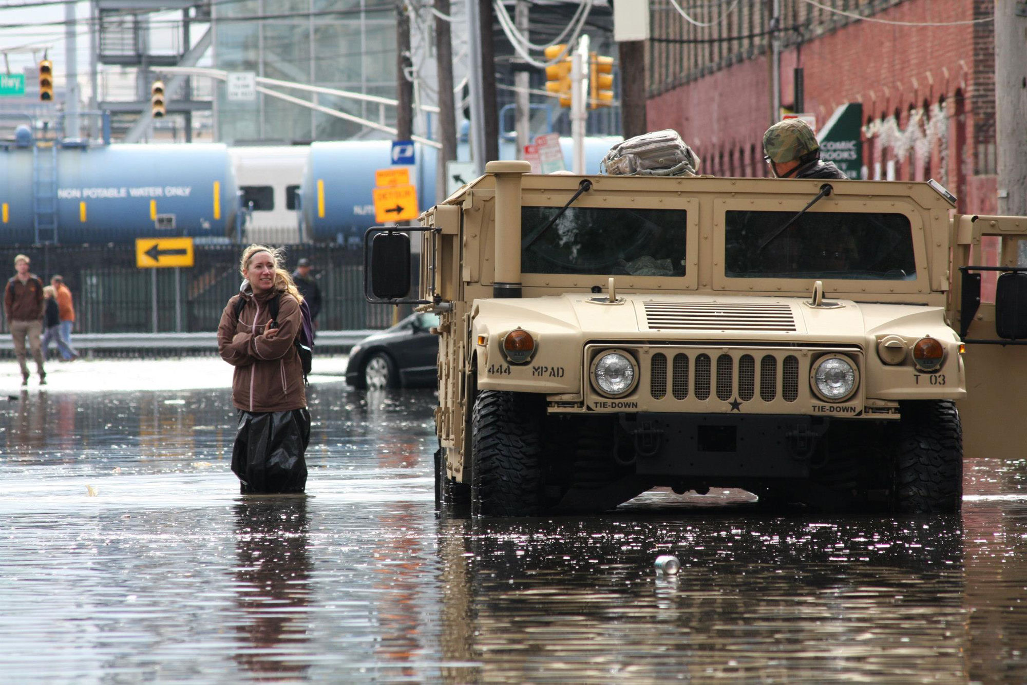 Photograph of flooding in the aftermath of Hurricane Sandy, Hoboken, New Jersey. The photo shows a beige humvee driving down a road coverd with shallow floodwaters. A woman stands to the left of the humvee in the water. She is wearing a brown coat and appears to have a plastic bag covering her legs. More people and part of a car can be seen in the background.
