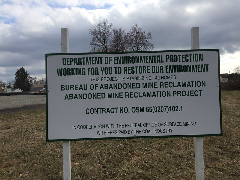 Photograph of a sign announcing a mine reclamation project in Pennsylvania. The sign says: "Department of Environmental Protection. Working for you to restore our environment. This project is stabilizing 142 homes. Bureau of Abandoned Mine Reclamation Abandoned Mine Reclamation Project. Contract No. OSM 65(0207)102.1. In cooperation with the Federal Office of Surface Mining with fees paid by the coal industry."
