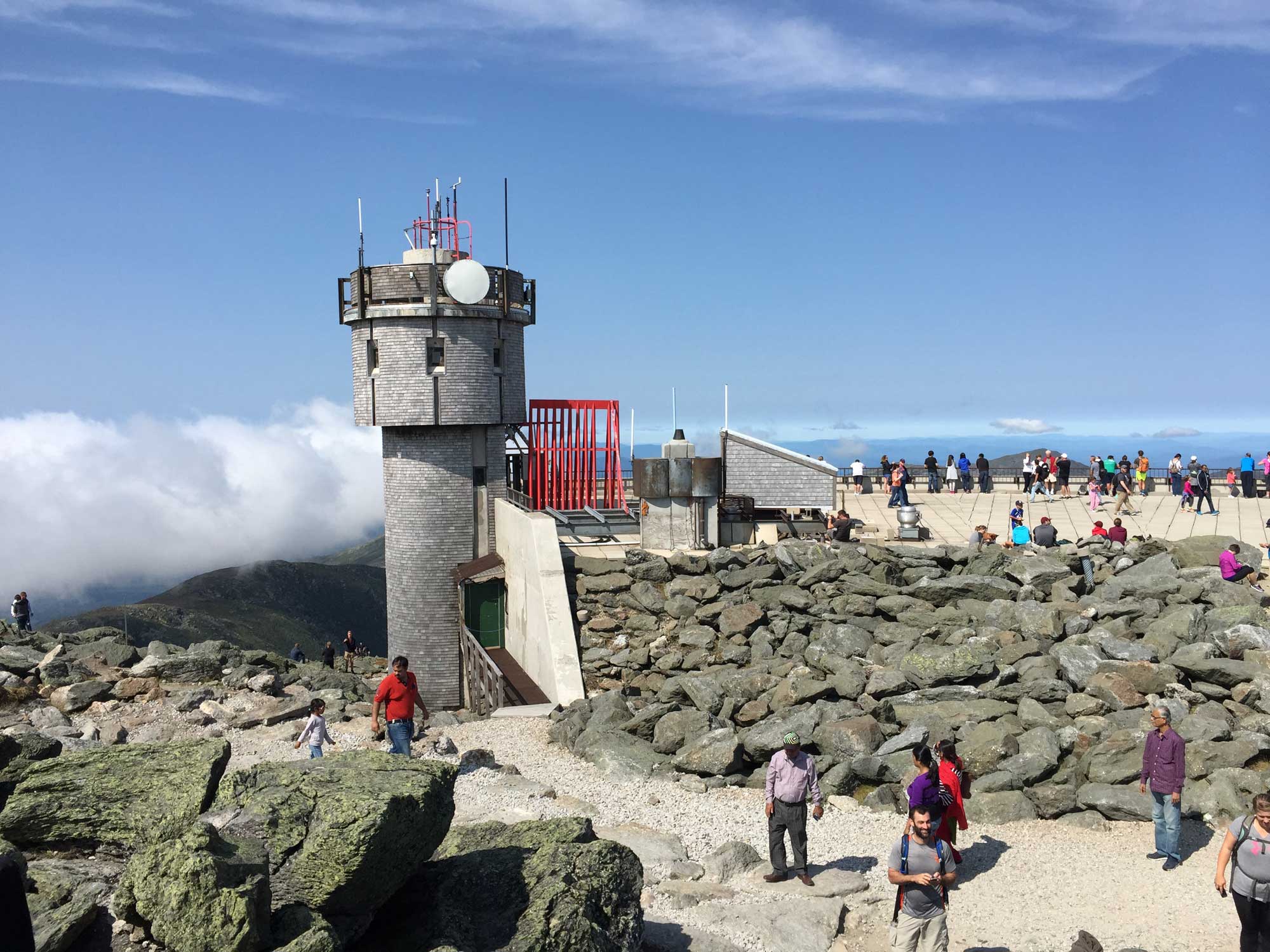 Photograph of the observation tower at Mount Washington Observatory in New Hampshire. The photo shows a cylindrical stone tower, trails, and a viewing area, with many people waking and standing.
