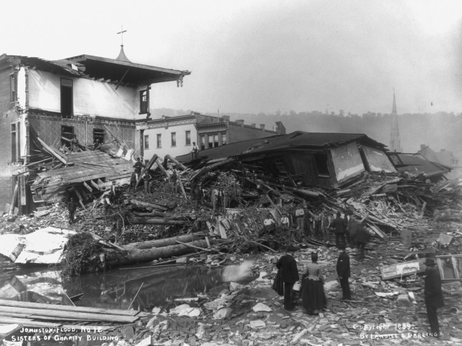 Black and white photograph showing the aftermath of the 1899 Johnstown Flood in Pennsylvania. The photo shows a collapsed building and debris, including a large uprooted tree. People and stand in the foreground looking at the wreckage, and some stand further back on the debris itself.