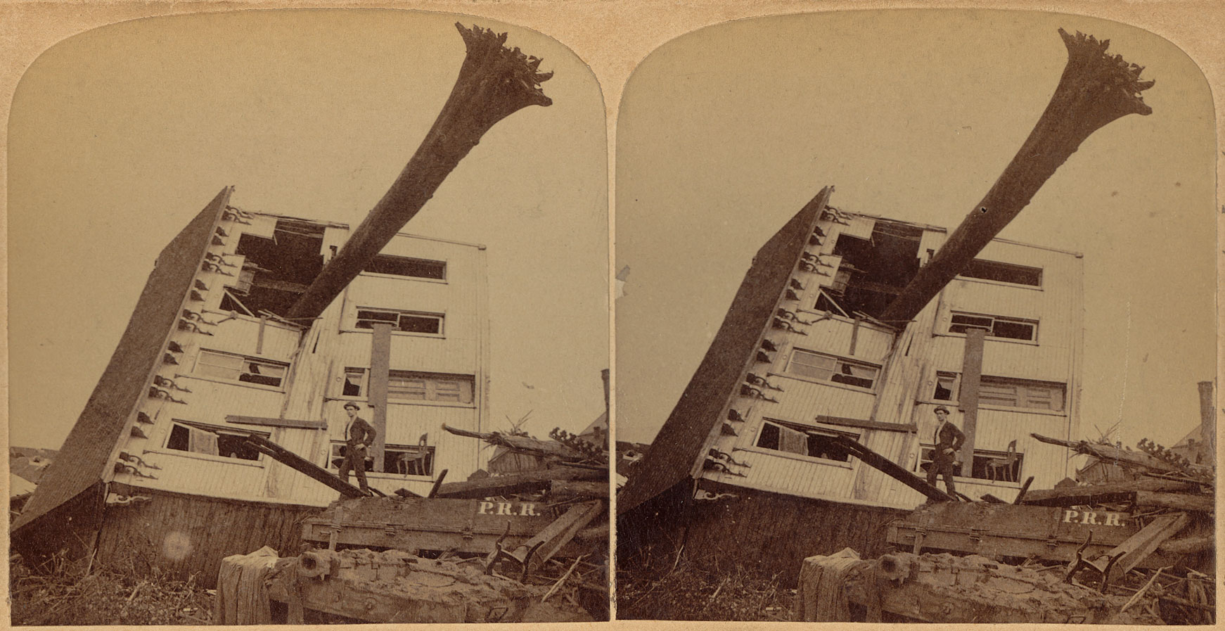 Stereopair (pair of nearly identical photographs placed side by side) showing a house destroyed by the 1899 Johnstown flood in Pennsylvania. The photo shows a house lying on its side with a large tree sticking out of one of the windows of the upper floor, the base of the trunk in the air. A man poses on a pile of debris in front of the house. The image is sepia.
