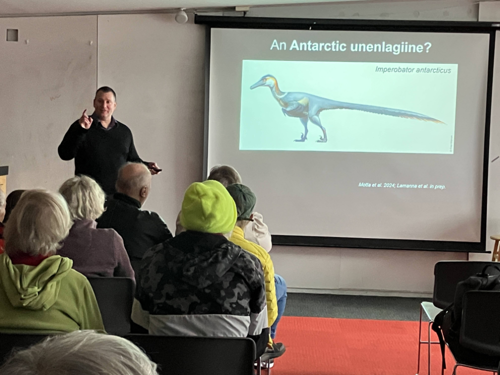 Photo of a man giving a lecture in a museum with a picture of a dinosaur